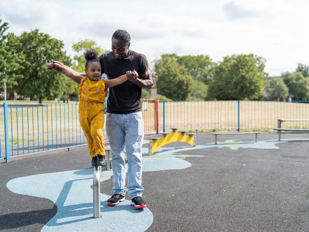 A dad guiding his daughter on a balance block at a playground